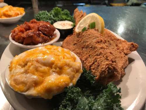 A plate of fried fish with lemon, served with cheesy mashed potatoes, a side of chicken, and garnished with kale.