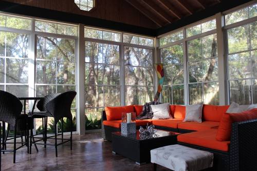 A bright sunroom with large windows, featuring an orange sofa, a coffee table, and a small dining area.