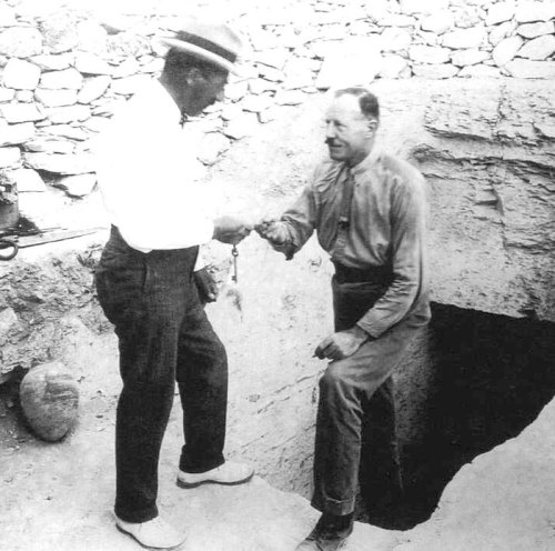 Two men shake hands near an excavation site, with a stone wall and a deep hole visible in the background.