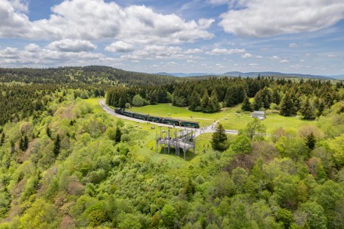 A scenic view of a train traveling through lush green hills and forests under a partly cloudy sky.