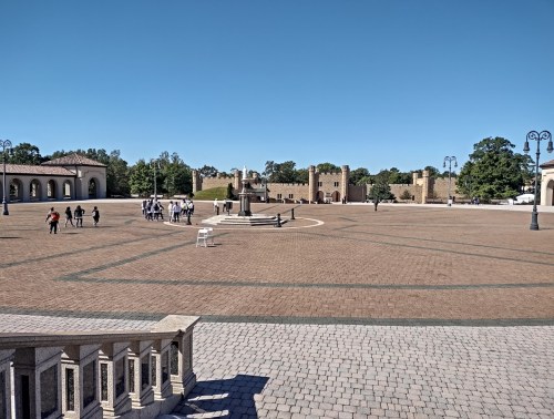 A spacious plaza with a statue at the center, surrounded by people and historic buildings under a clear blue sky.