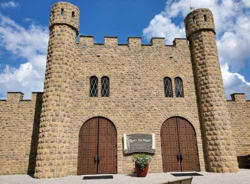 Stone castle entrance with two towers, large wooden doors, and a sign in front against a blue sky with clouds.