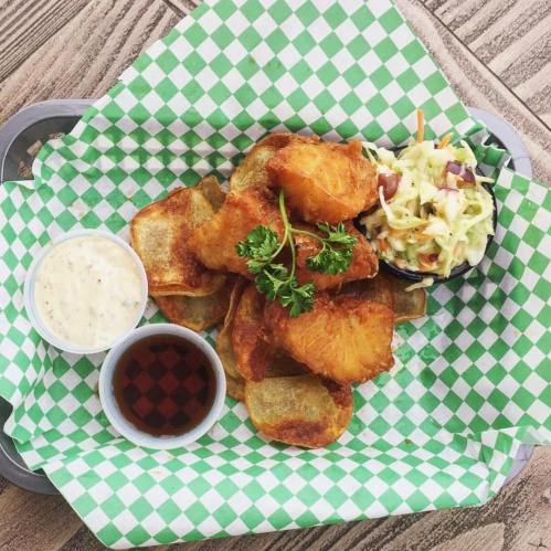 A plate of crispy fried fish with chips, coleslaw, and dipping sauces on a green checkered paper.