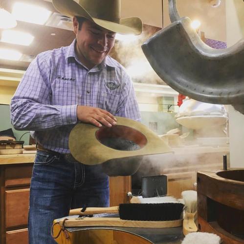 A man in a cowboy hat shapes a hat in a workshop, surrounded by tools and steam.