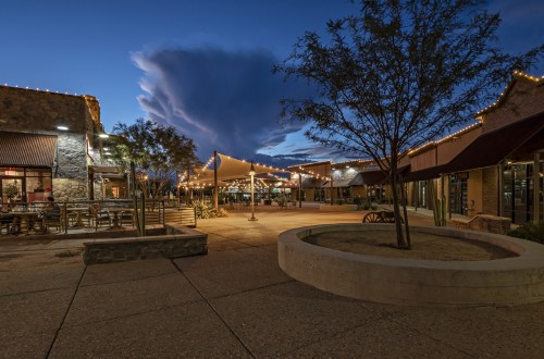 A charming outdoor shopping area at dusk, with string lights, a large cloud, and a circular seating area.