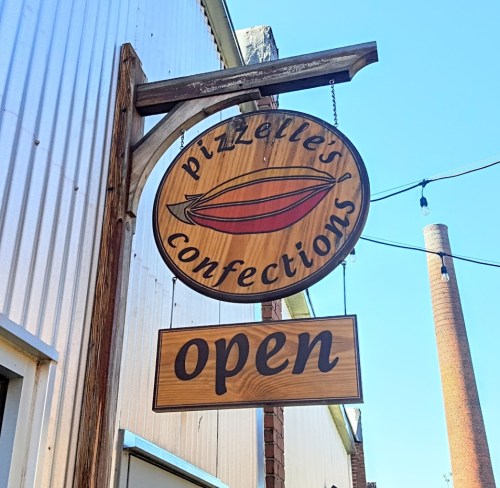 Sign for Pizzelle's Confections, featuring a boat graphic and "Open" below, against a clear blue sky.