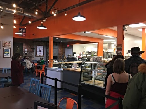 A cozy café interior with colorful seating, a display case, and patrons waiting in line. Warm lighting and brick walls.