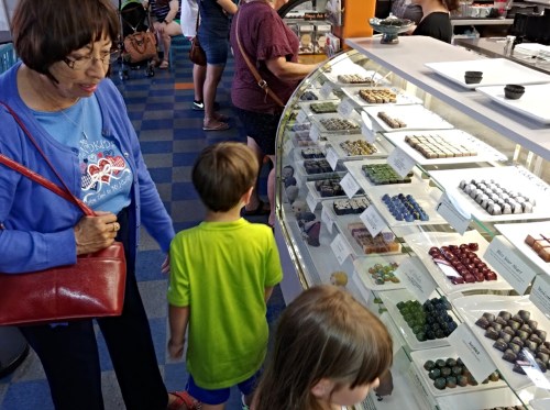 A woman and two children look at a display of colorful chocolates in a shop, with other customers in the background.