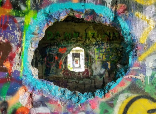 A colorful graffiti-covered wall with a large hole revealing a bright window in the distance.