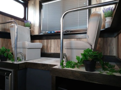 Interior of a cozy space featuring two white chairs, plants, and wooden accents, with natural light coming through a window.