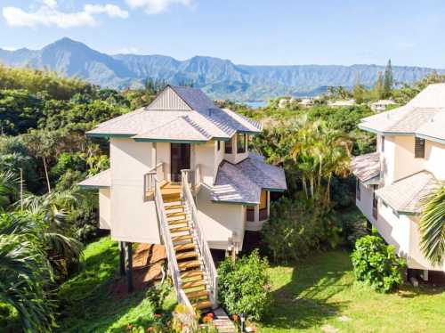 A two-story house with a staircase, surrounded by lush greenery and mountains in the background under a clear blue sky.