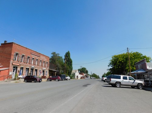 A quiet small town street with brick buildings, parked cars, and clear blue skies.