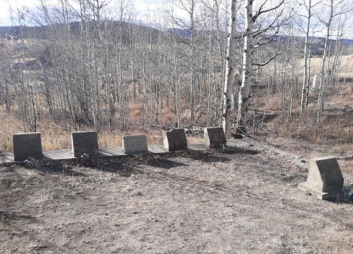 A row of simple stone grave markers in a sparse, wooded area with dry grass and distant hills.