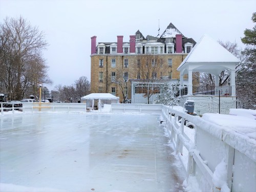 A snowy outdoor ice skating rink with a historic building in the background, surrounded by bare trees.