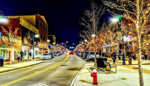 A vibrant street at night, lined with trees adorned with lights, shops, and parked cars, creating a festive atmosphere.