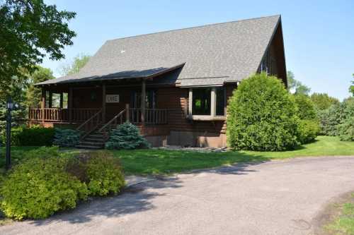 A rustic wooden house with a sloped roof, surrounded by greenery and a gravel driveway on a sunny day.