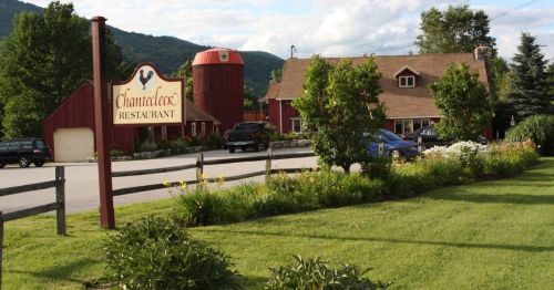 A charming restaurant with a red barn, surrounded by greenery and flowers, set against a mountainous backdrop.