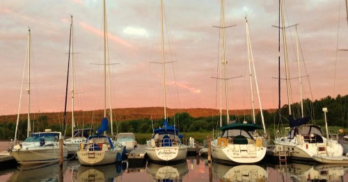 Sailboats docked at a marina during sunset, with a pink sky and lush greenery in the background.