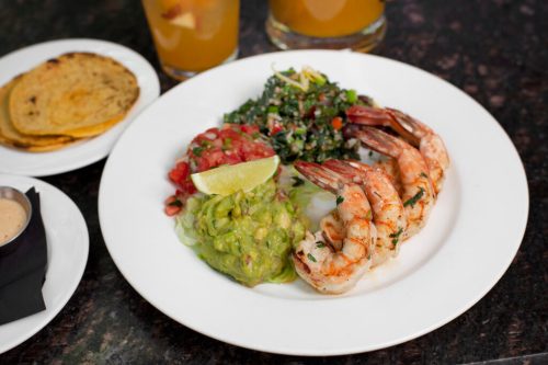 A plate of grilled shrimp with guacamole, pico de gallo, and a side salad, accompanied by tortillas and drinks.