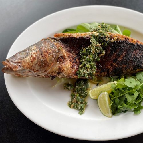A whole grilled fish topped with green sauce, served on a white plate with lime wedges and fresh herbs.