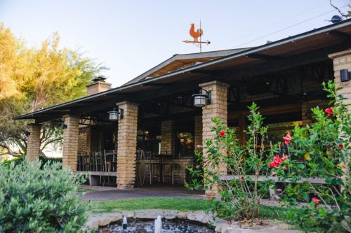 A rustic restaurant with stone pillars, outdoor seating, and a weather vane, surrounded by greenery and flowers.