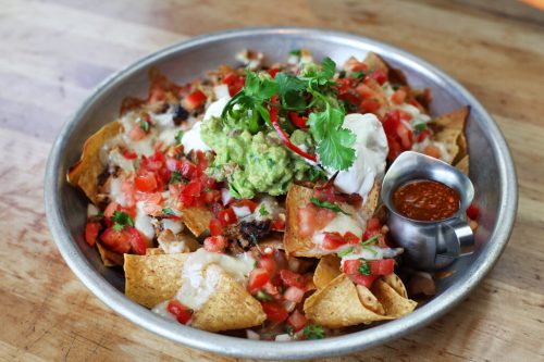 A platter of nachos topped with cheese, guacamole, sour cream, diced tomatoes, and a side of salsa.