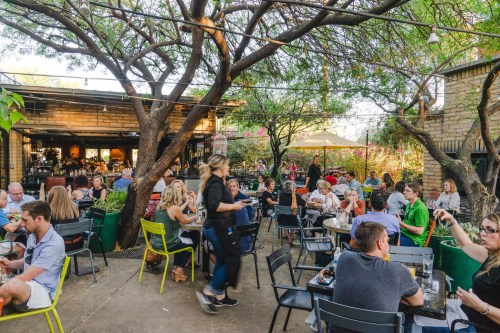 A lively outdoor restaurant scene with diners at tables under trees, enjoying food and drinks in a vibrant atmosphere.