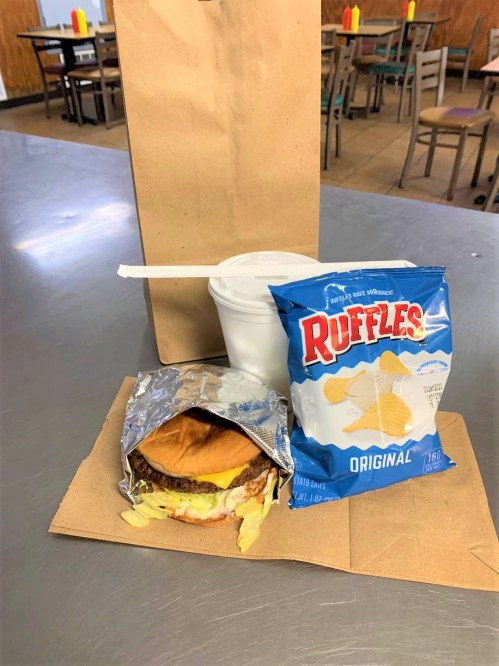A paper bag holds a burger with cheese, a cup, and a bag of Ruffles chips on a table in a casual dining setting.