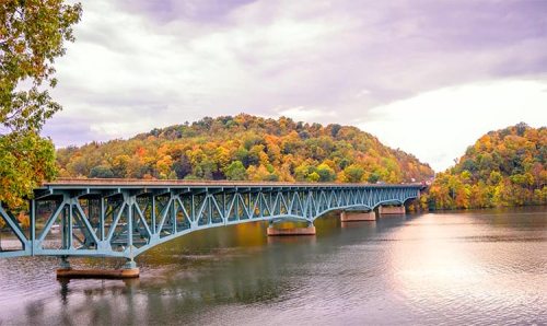 A blue bridge spans a calm river, surrounded by vibrant autumn foliage on rolling hills under a cloudy sky.