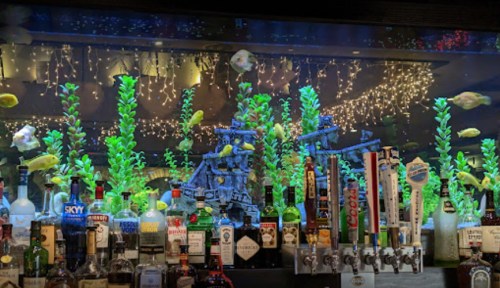 A bar with a colorful aquarium backdrop featuring fish, plants, and a ship model, illuminated by soft lights.
