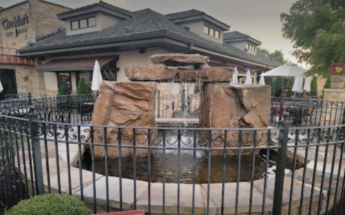 A stone fountain with water flowing, surrounded by a black fence and outdoor seating at a restaurant.