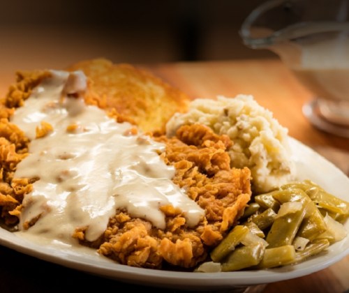 A plate of fried chicken topped with gravy, served with green beans, mashed potatoes, and cornbread.