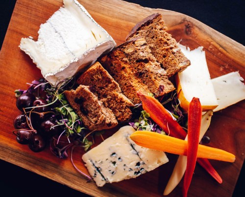 A wooden platter featuring assorted cheeses, bread, grapes, and colorful carrots, garnished with microgreens.