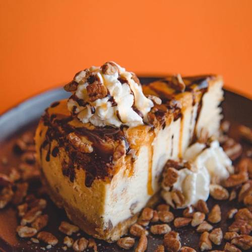 A slice of cheesecake topped with whipped cream, chocolate drizzle, and crushed nuts on a dark plate against an orange background.