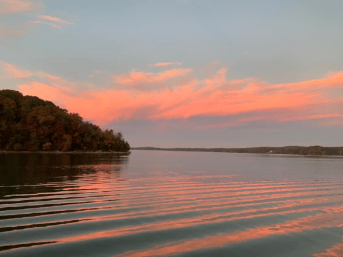 A serene lake at sunset, with colorful clouds reflecting on the water and a wooded shoreline in the background.