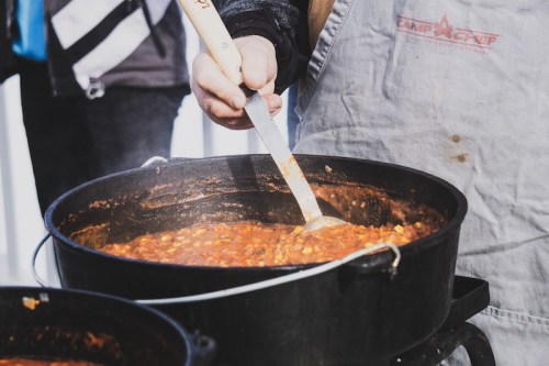 A person stirs a pot of chili over a campfire, wearing an apron and holding a wooden spoon. Steam rises from the pot.