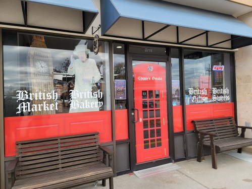 Exterior of a British-themed shop with red doors, featuring signs for a market, bakery, gifts, and sweets.