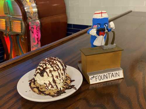 A dessert with whipped cream and chocolate drizzle next to a playful figurine of a soda fountain.