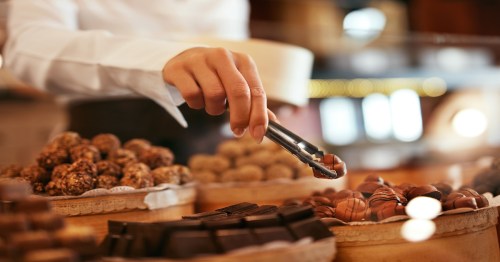 Chocolate Candies In Confectionery Store. Closeup Of Female Hand With Forceps Choosing Candy