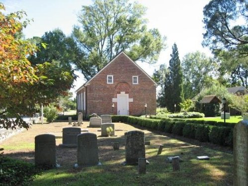 A peaceful cemetery with gravestones in front of a brick church, surrounded by trees and greenery.