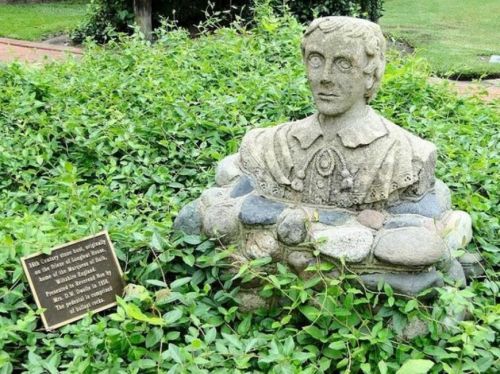 A stone bust of a man surrounded by greenery, with a plaque nearby detailing its significance.