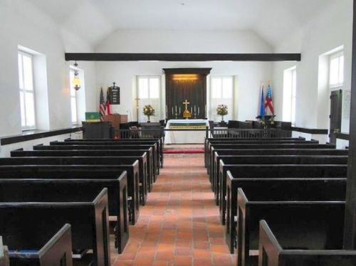 Interior of a simple church with wooden pews, an altar, and flags, featuring a bright, airy atmosphere.