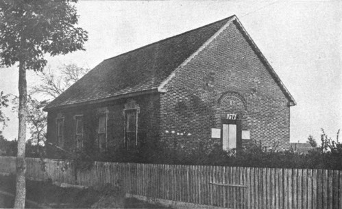Historic brick building with a gabled roof, surrounded by a wooden fence and trees, in a rural setting.