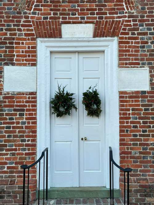 White double doors with green wreaths, set against a brick wall, featuring a stone inscription above.