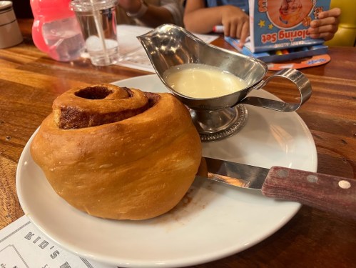 A cinnamon roll on a plate with a small silver gravy boat of cream beside it, set on a wooden table.