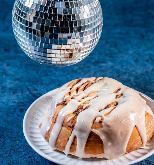 A glazed cinnamon roll on a plate, with a disco ball hanging above it against a blue background.