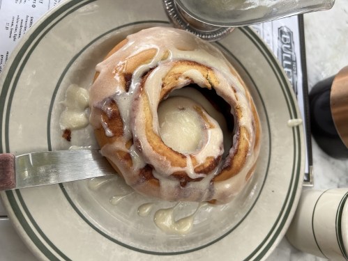 A freshly baked cinnamon roll topped with creamy icing, served on a plate with a knife beside it.