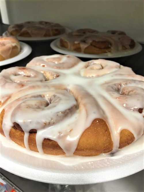 A close-up of a glazed cinnamon roll on a white plate, with several more rolls in the background.