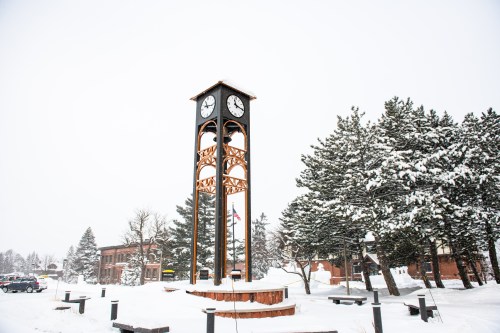A tall clock tower surrounded by snow-covered trees and a winter landscape, with light snowfall in the scene.