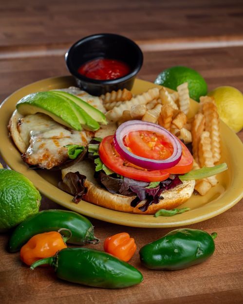 A colorful burger with lettuce, tomato, onion, and avocado, served with crinkle fries and dipping sauce on a wooden table.